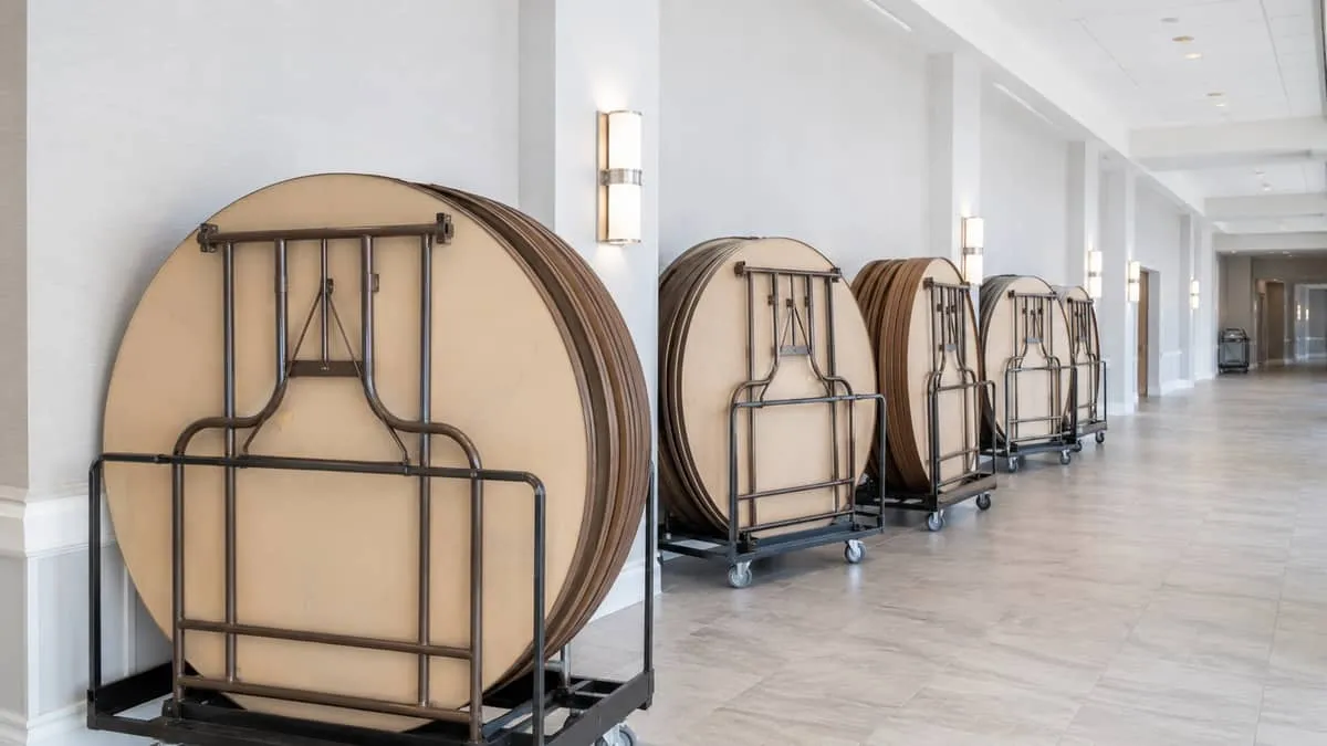 a row of 60-inch round folding banquet tables on rolling storage carts in a hotel back-of-house corridor, showing the pr