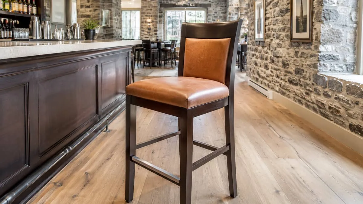 close-up of a commercial barstool with a dark wood frame and cognac leather seat, positioned against a stone wall interi