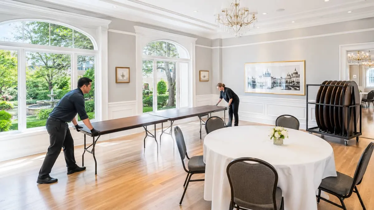 a Victoria event venue mid-transition, round tables being swapped for long banquet tables by two staff members, folding 